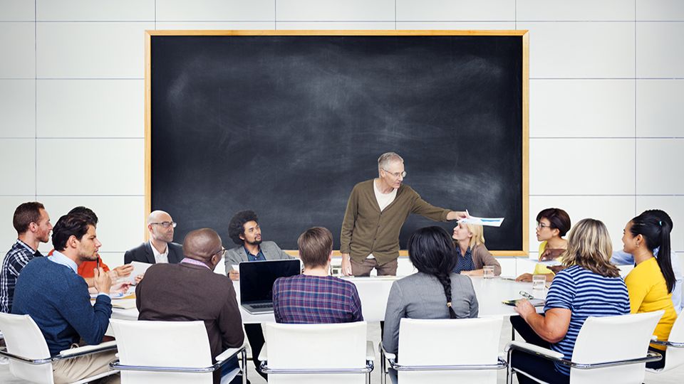 A grey haired man stands in front of a large blackboard and addresses a multiethnic group of students. The students are sitting around a large table.