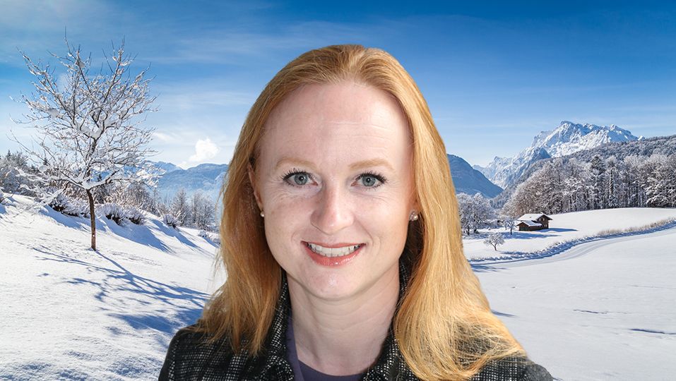 Caroline Langley against a backdrop of a beautiful winter scenery with trees and mountain tops in the Alps on a sunny day with blue sky and clouds.