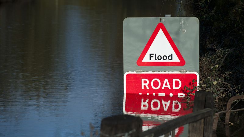 Rising flood water partially submerges a warning sign advising motorists that the road/highway is closed.