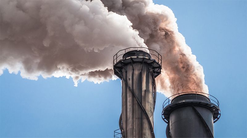 Close-up showing the top of two large chimneys emitting thick clouds of smoke into a clear blue sky.