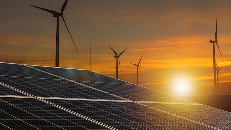 Illuminate your project with this stunning image of renewable energy synergy, capturing solar panels in the foreground with wind turbines silhouetted against the vibrant hues of a setting sun.