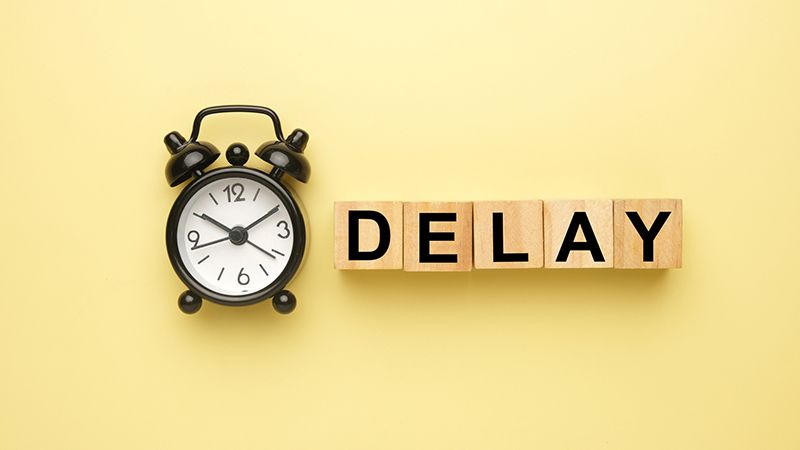 Flatlay picture of alarm clock with wooden block written delay on yellow background.