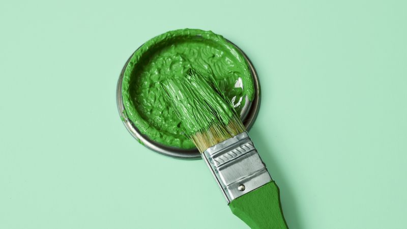 closeup of a paintbrush and the lid of a paint can with green pain, on a pale green background